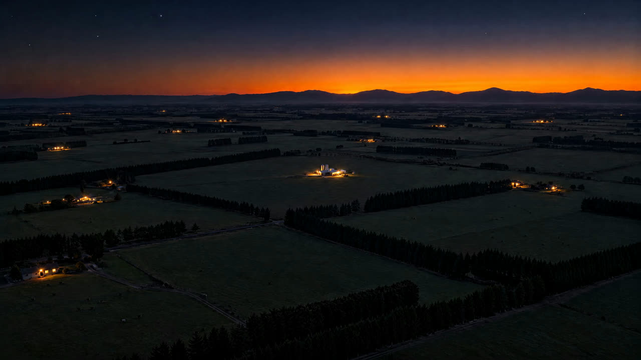 Dark dairy plains before dawn, farm lights scattered under an orange band of light along the hills